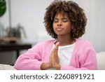 Small photo of Stress relief, mediation, mind fullness concept. Closeup of peaceful millennial black woman with bushy hair in homewear sitting alone on couch at home, holding hands in namaste gesture, copy space