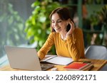 Cheerful beautiuful brunette young woman in smart casual entrepreneur attending online business meeting, woman sit at table in front of laptop, using earpods, taking notes, cafe interior, copy space