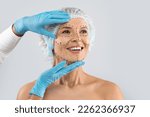 Small photo of Cheerful excited middle aged woman in medical hat with pre surgery marks on her face looking at copy space for ad and smiling, surgeon hands in blue gloves touching female skin