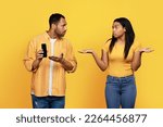 Small photo of Sad serious young african american guy show phone to confused woman isolated on yellow background, studio. Jealousy, relationship and love problems, gadget addiction, social network and online mistake