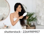 Happy millennial caucasian brunette lady in towel combing long curly hair, enjoy spa treatment in bedroom interior. Routine procedures at home and good morning, beauty care at weekend and spare time