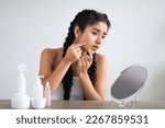 Small photo of Skin Problems. Upset Young Indian Woman Looking At Pimple On Her Cheek, Sad Beautiful Hindu Female Sitting Near Magnifying Mirror At Home And Inspecting Her Face, Suffering Acne, Copy Space
