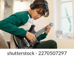 Asian Teen Guy Playing Electric Guitar Wearing Headphones Writing Song Sitting At Home. Boy Learning To Play Musical Instrument On Weekend. Teenager's Hobby And Music Talent. Selective Focus