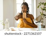 Funny black woman covered in towel enjoying morning routine, singing and dancing with hairbrush, having fun after shower, sitting in bedroom interior, free space