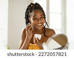 Portrait of beautiful black woman applying facial cream and smiling, lady moisturizing skin with lifting nourishing day creme after morning shower