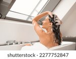 Haircare Cosmetics. Lady Applying Shampoo On Long Hair Washing Head Sitting In Bathtub Full Of Foam Indoors. Side View Shot Of Happy Woman Enjoying Hair Care Routine In Modern Bathroom