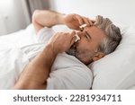 Exhausted sick middle aged grey-haired bearded man wearing pajamas lying in bed with fever and runny nose, touching head and sneezing nose, suffering from cold, flu, coronavirus, closeup