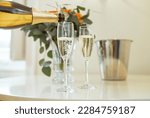 Closeup Of Sparkling Wine Bottle And Alcohol Pouring In Two Champagne Glasses Standing On Table Near Bucket And Vase With Flowers Indoors. Drinks Serving Concept. Cropped Shot, Selective Focus