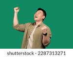 Emotional asian man shouting and shaking fists with eyes closed, celebrating big success over green studio background. Celebration of victory, joy emotion concept