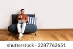 Small photo of Relax concept. Happy asian man drinking coffee using pc sitting on comfortable sofa and dreaming, panorama with free space. Male resting on sofa, enjoying weekend free time