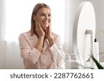 Beautiful Middle Aged Woman Touching Face And Looking In Mirror At Home, Happy Mature Attractive Lady Enjoying Her Flawless Skin, Smiling Female Making Anti-Aging Beauty Routine, Closeup