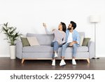 Small photo of Excited cheerful happy young indian woman and man in casual sitting on couch at home, using the remote to turn on the ac unit to cool down during the hot weather, pointing at copy space