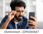 Small photo of Eyesight Problems Concept. Young Indian Man In Eyeglasses Looking At Smartphone Screen And Squinting, Eastern Guy Trying To Read Message, Suffering From Astigmatism And Bad Vision, Closeup Shot