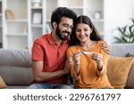 Portrait Of Happy Young Indian Couple Holding Positive Pregnancy Test While Sitting On Couch At Home Together, Eastern Husband Embracing His Joyful Wife, Celebrating Awaiting Baby, Copy Space