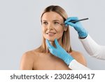 Beautician hands wearing blue medical gloves drawing contours with black pen on woman face before beauty treatment, middle aged blonde lady getting beauty injections, isolated on grey, copy space