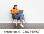 Happy pretty young long-haired arab woman in casual outfit sitting in armchair over white wall background, typing on pc laptop keyboard and smiling, female digital nomad working from home, copy space