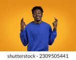 Small photo of Making Wish. Superstitious emotional stylish black guy in blue sweater with closed eyes keeping his fingers crossed, praying for luck, isolated on yellow studio background, copy space