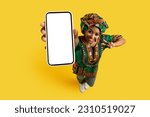 Smartphone with white blank screen in pretty black woman hand, happy young lady wearing national african costume showing great online deal and peace gesture, yellow studio background, mockup, top view