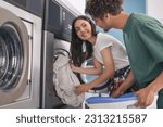 Happy Couple Folding Laundry Together At Laundromat Washing Machine Indoor, Smiling To Each Other Enjoying Convenience Service Together On Weekend. Spouses Washing Clothes At Public Laundrette
