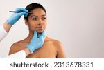 Happy attractive young black woman attending aesthetic clinic, unrecognizable doctor plastic surgeon making pre surgery marks on pretty lady face before beauty procedure, copy space, web-banner