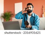 Small photo of Financial Hardship. Unhappy Young Indian Man At Laptop Holding Empty Wallet, Counting Money Struggling Finance Problems Sitting On Couch At Home. Guy Suffering From Economic Crisis