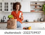Small photo of Shopping Economy. Happy Black Woman In Kitchen Checking Bill After Grocery Shopping, Smiling African American Female Standing Near Table, Counting Spends, Enjoying Affordable Prices For Food
