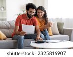 Happy Indian Spouses Checking Financial Documents And Calculating Family Budget At Home, Cheerful Eastern Couple Sitting At Table In Living Room, Reading Loan Papers Or Counting Monthly Expenses