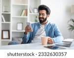 Smiling Young Indian Man Using Smartphone And Drinking Coffee At Home, Happy Millennial Eastern Guy Browsing Social Networks On Mobile Phone While Relaxing At Desk Indoors, Copy Space