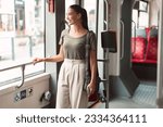 Happy Passenger Lady Standing In Modern Tram Enjoying Comfortable Ride In Public Transport Indoor, Looking Away Out Of Window. Student Commuting To University By Bus Or Trolleybus