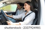Small photo of Cheerful confident pretty young woman sitting inside car with man instructor by her side, lady student holding hands on steering wheel, passing exams successfully at driving school, getting licence