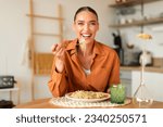 Happy young european lady eating delicious homemade Italian pasta, enjoying tasty lunch, looking at camera and laughing, sitting in cozy kitchen interior, copy space