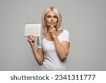 Small photo of Pensive Mature Lady Holding Blank Menstrual Calendar And Touching Chin, Thoughtful Senior Female Suffering Lack Of Menstruation, Having Climacteric Symptoms Or Menopause, Standing On Grey Background