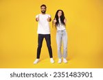 Happy arab millennial man and woman in casual showing thumb ups and smiling at camera, posing full length, showing like gestures with both hands standing over yellow background, studio shot