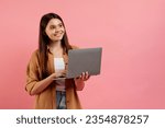 Small photo of Beautiful smiling teen girl holding laptop and looking aside at copy space, young female student using computer for online education and distance learning, standing on pink background, copy space