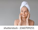 Beauty and skincare concept. Portrait of beautiful mature woman with towel on head touching her face, looking aside at free space and smiling, grey background