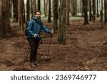 Glad young caucasian man in jacket with trekking sticks walk, enjoy adventure at free time at weekend in cold forest in autumn. Travel, vacation together, active lifestyle, full length