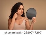 Small photo of Upset brunette young woman wearing beige top looking at mirror and touching her face over beige studio background, suffering from skin problems. Acne, wrinkles, dull skin, early premature aging