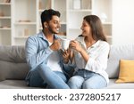 Small photo of Cheerful young indian couple enjoying leisurely moment on sofa, drinking coffee while having pleasant conversation, creating heartwarming, homey scenario