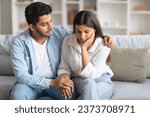 Small photo of Caring young indian husband next to wife, lovingly attempting to soothe and console woman, providing comforting presence, sitting on sofa at home