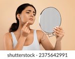 Small photo of Skin Problems Concept. Unhappy Young Indian Woman Holding Magnifying Mirror And Looking At Pimple On Her Chin, Upset Eastern Female Inspecting Acne, Standing Isolated Over Beige Studio Background