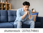 Small photo of Depression And Mental Struggle. Asian young man sitting expressing sad emotion, having depression and stress symptoms on sofa in living room interior. Emotional hardship and unhappiness