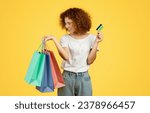 Small photo of Cheerful young european woman shopaholic, with colorful bags, credit card, enjoy shopping, isolated on yellow background, studio. Shopaholic with purchases, recommends money for sale