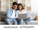 Small photo of Indian young couple, comfortably settled on sofa in homey setting, shares a delightful moment browsing through laptop, with shelves filled with books in the background