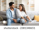 Small photo of Excited young Indian couple enjoys light-hearted moment, sitting close together on sofa, sharing laughter and warmth, looking at each other and smiling
