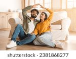 Small photo of Young couple playfully forming symbolic roof or house shape with their hands, sitting together on floor near couch at home. Both have joyous expressions, signifying comfort and happiness