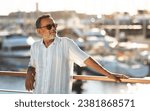 Small photo of Escapade. Relaxed senior man with sunglasses, standing near luxurious yachts at the dockside, enjoys his summer holiday in marina, looking aside. Seaside relaxation and adventurous retirement