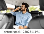 Small photo of Always In Touch Concept. Young indian guy manager in glasses writing in notebook talking on smart phone sitting in back seat of car, looking through window, taking taxi