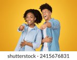 Small photo of Laughing heartily, a young african american woman and a young man stand close to each other, pointing at the camera as if sharing a joke, set against a cheerful yellow background