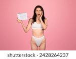 Small photo of Shocked glad young caucasian woman in white underwear holding a calendar with a surprised expression, symbolizing forgotten events or deadlines on a pink background. Wellness, body care lifestyle