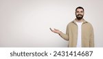 Small photo of Affable bearded man smiling and gesturing with his hand open as if holding something invisible, dressed in a casual beige shirt and white t-shirt, on a wide light background